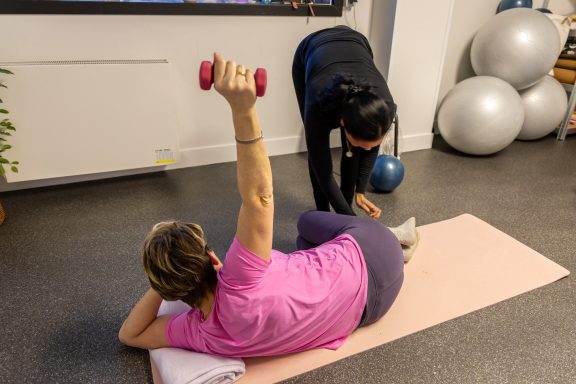 Vrouw ligt op een pilatesmat en tilt een dumbbell op, terwijl Nazrien haar begeleidt.