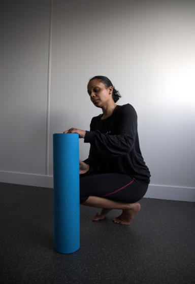 Vrouw in zwarte kleding hurkt naast een blauwe pilates mat, in een lichte ruimte.