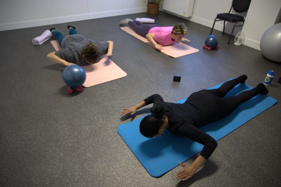 Nazrien en twee deelnemers doen pilates oefeningen op pilatesmatten met gewichten en een gymbal.