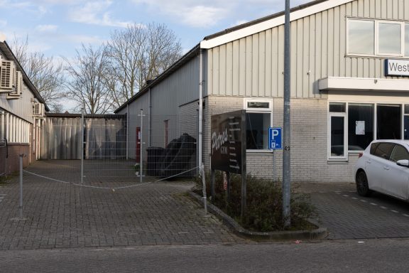 Bedrijfsgebouwen met een parkeerplaats. Achter het hekwerk ligt de ingang van de pilates studio.